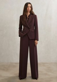 Dviejų eilučių pritaikytas kostiumas gilia bordo spalva, su platėjantiems kojų raukšlėmis ir tvarkingomis, struktūruotomis pečių linijomis, pagamintas iš lygios medžiagos.