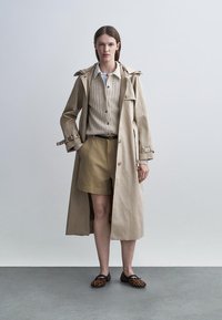 Woman standing wearing beige trench coat, ribbed beige cardigan, tan shorts, and leopard print flats on a gray floor with plain background.