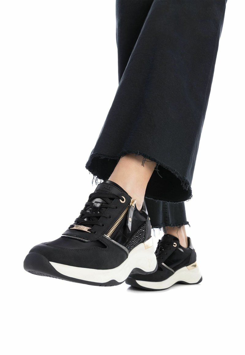 Zapatillas negras con un diseño texturizado, que presentan un detalle con cremallera y acentos dorados. Suela blanca gruesa y los puños de los pantalones negros parcialmente deshilachados.