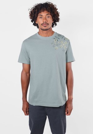 Homme aux cheveux bouclés portant un t-shirt vert clair avec un imprimé floral sur l'épaule et un pantalon sombre, debout face à l'avant sur un fond uni.