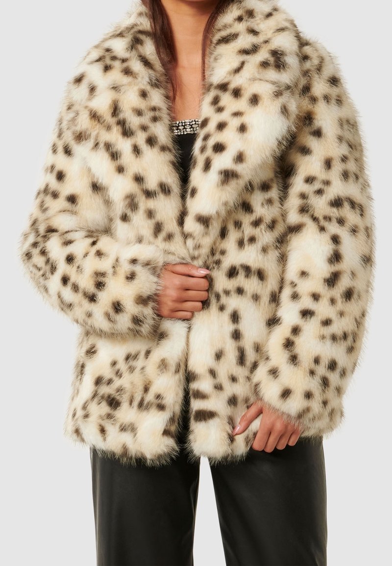 Femme portant un manteau en fausse fourrure beige avec des taches de léopard brun foncé, assorti à un pantalon noir et un haut noir orné de perles en-dessous.