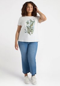 Blusa corta de manga corta blanca con diseño abstracto de hojas en verde y amarillo; combinada con pantalones vaqueros de azul claro y zapatillas blancas.