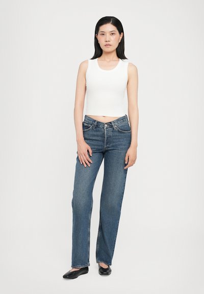 Débardeur blanc côtelé associé à un jean droit bleu taille haute. Le jean dispose d'une fermeture à bouton et à zip classique, avec peu d'usure.