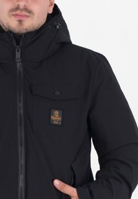 Zwarte jas met ritsluiting, verstelbare capuchon en een borstzak met knoop en een logo-opdruk. Gladde, duurzame stof.