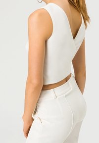 Donna che indossa un top corto bianco senza maniche con una schiena a forma di V e pantaloni bianchi a vita alta su uno sfondo neutro.