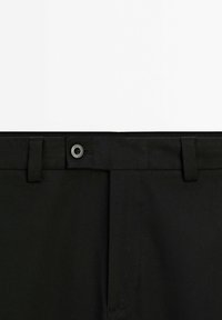 Pantaloni neri con una cintura dotata di passanti per cintura, pieghe frontali e chiusura con bottone. Il tessuto presenta un motivo testurizzato sottile.