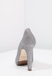 Stříbrná třpytivá vysoká bota viděná z zadní strany, stojící na bílé ploše na bílém pozadí.