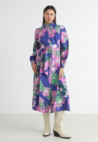 Blommig klänning med livfulla rosa, gröna och blå färger. Långa ärmar, hög hals, bältad midja och knäppning med en svävande silhuett.