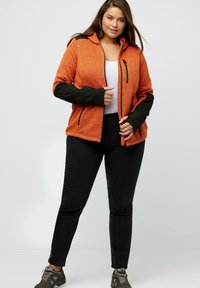 Giacca in fleece arancione con dettagli neri, tasche con zip e cappuccio, abbinata a pantaloni neri e scarpe da trekking grigie. Texture liscia.