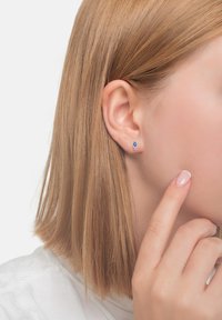 Silvriga studs med en blå och rosa ädelsten i tårform, bärs i ett öra, med ljust brunt hår som är stylat rakt.