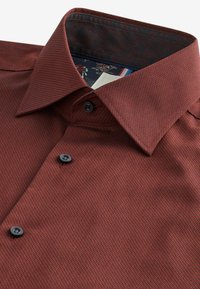 Chemise texturée bordeaux avec un col doux, ornée de boutons noirs et d'une doublure intérieure à motifs. Le tissu présente un motif à carreaux subtil.
