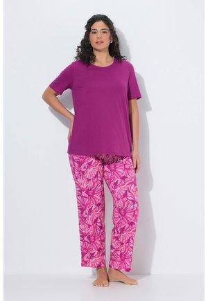 Kvinde, der står barfodet iført en ensfarvet lilla kortærmet skjorte og løstsiddende bukser med lyserødt blomsterprint mod en hvid baggrund.