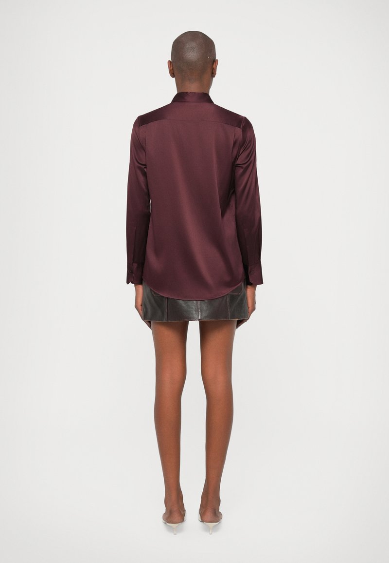 Eine Person steht mit dem Rücken zu uns und trägt eine langärmelige, dunkelburgunderfarbene Bluse, einen schwarzen, kurzen Lederrock und weiße High Heels vor einem neutralen Hintergrund.