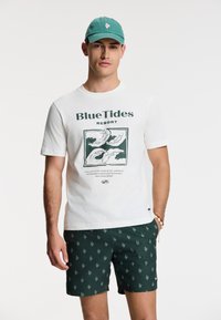 Férfi zöld sapkában, fehér "Blue Tides Resort" feliratú pólóban, hullámos mintás zöld shortban és kagyló karkötővel, zsebébe tett kézzel áll.