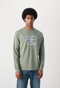 Hosszú ujjú olívazöld pamut póló, amelyen elöl egy világoskék grafikai tervezés látható: "Makia Clothing Co.". Szabványos szabású, kerek nyakkivágással.