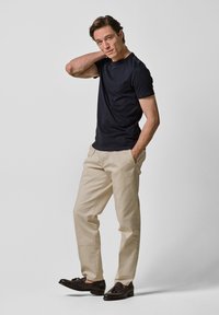 Marinblå kortärmad t-shirt, beige byxor och mörka skor. Modellen poserar med en hand på nacken och visar upp en avslappnad stil och passform.