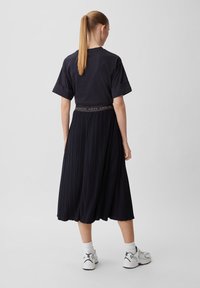 Donna con coda di cavallo, che indossa una maglietta nera oversized, una gonna midi plissettata nera con cintura con logo, calzini bianchi e sneakers bianche, vista di spalle.