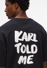 Melna kokvilnas t-kreklu ar izteiksmīgiem baltiem burtiem uz muguras, kas lasa "KARLS MAN TEICA," ar brīvu piegriezumu un īsām piedurknēm.