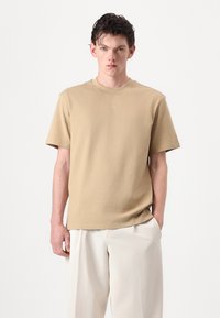 Beige bomulds t-shirt med afslappet pasform og korte ærmer. Har en rund halsudskæring og en diskret logo detalje i midten.