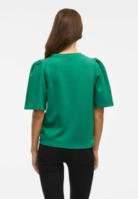 Top verde de manga corta con hombros abullonados, escote redondeado y tejido suave. Vista desde la parte posterior, combinado con pantalones negros.