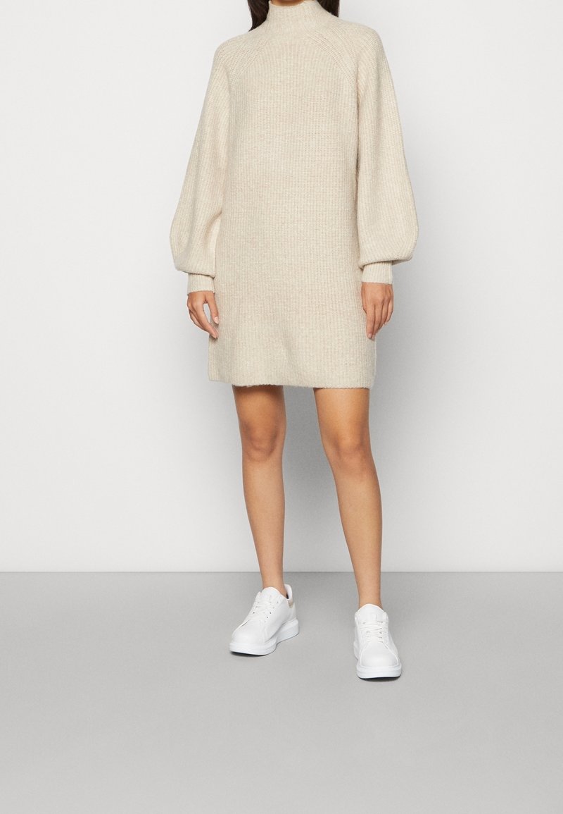 Robe pull tricotée crème avec un col montant, des épaules tombantes et des manches bouffantes, présentant une coupe décontractée et arrivant à mi-cuisse.