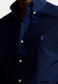 Dunkelblaue Button-up-Bluse aus Baumwollstoff mit klassischem Kragen, kontrastierenden hellen Knöpfen und einem gestickten Logo auf der Brust.