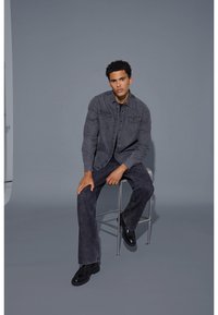 Grå denimskjorta med två bröstfickor, kombinerad med matchande mörka jeans och svarta skor, sittande på en pall mot en neutral bakgrund.