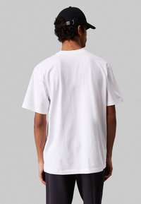 Maglietta bianca in cotone con maniche corte, colletto a giro, vestibilità comoda e una texture liscia. Cappellino nero indossato sulla testa del modello.