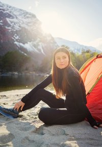 Sort termisk trøje og leggings båret af en person, der sidder på sand, iført blå vandresko. Et orange telt er opsat i nærheden foran en bjerglandskab.