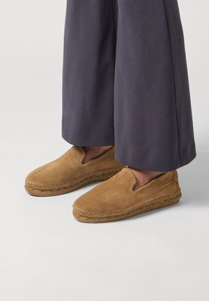 Braune Wildleder-Espadrilles zum Hineinschlüpfen mit geflochtenen Jutesohlen, getragen zu dunklen, weit geschnittenen Hosen.