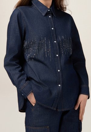 Kobieta w ciemnej koszuli z denimu zapinanej na guziki, ozdobionej pionowym wzorem ze srebrnych ćwieków na wysokości klatki piersiowej, oraz w dopasowanych spodniach z denimu, z rękami w kieszeniach.