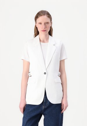 Blazer sin mangas blanco con cuello con muescas y cierre de un solo botón. Presenta aros laterales y bolsillos frontales. Llevado sobre una camiseta blanca.