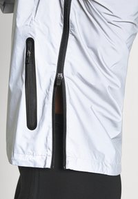 Jasnoszara kurtka z czarnymi zamkami błyskawicznymi. Posiada przedni zamek oraz boczną kieszeń zapinaną na zamek. Gładka faktura, lekki materiał.