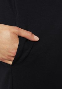Musta kangas, jossa on sileä pinta, sisältää osittain peitetyn sivutaskun, johon on asetettu käsi. Yksinkertainen muotoilu, jossa on toiminnallisia yksityiskohtia.
