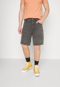 Pantalones cortos de mezclilla negra con corte recto, con bolsillos frontales y un aspecto deslavado, combinados con zapatillas altas amarillas y una camisa a rayas naranja.
