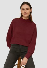 Bordeaux gebreide trui met een hoge kraag, lange mouwen en geribbelde manchetten. Gekozen in combinatie met donkere jeans, zittend op een houten kruk tegen een grijze achtergrond.