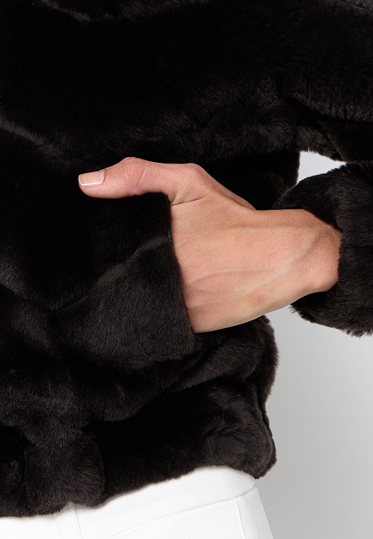 Schwarze Kunstfelljacke mit plüschiger Textur und einer seitlichen Tasche, in die eine Hand gesteckt ist, um das Kleidungsstück zu demonstrieren. Einfaches, weiches Design.