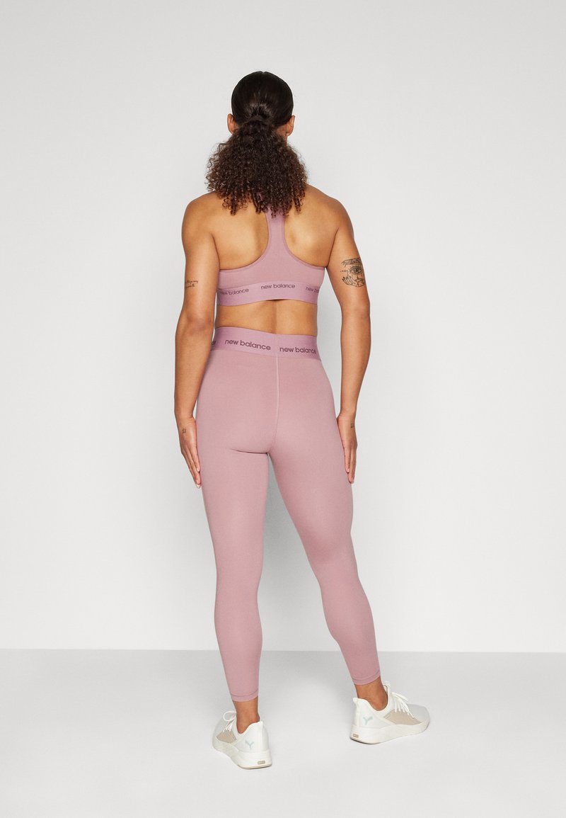 Mujer de pie de espaldas con sujetador deportivo y mallas color malva de New Balance y zapatillas blancas sobre fondo liso.