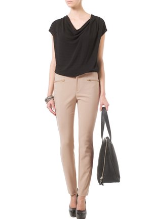 Woman wearing a black draped top, beige slim pants with zipped pockets, black heels, bracelet, holding a large black handbag by the straps.
