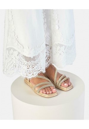 Pies con sandalias beige de tiras y dedos cuadrados pequeños, de pie sobre un pedestal redondo blanco, con un vestido blanco con encaje en el borde.