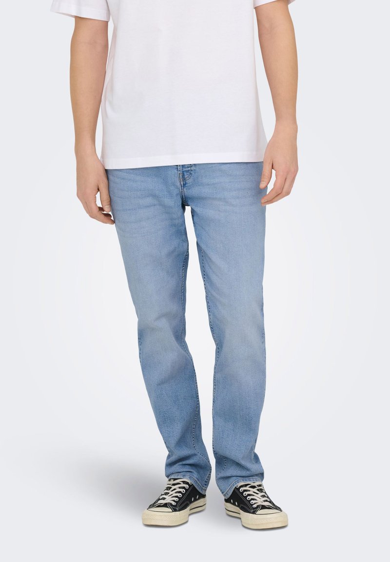 Personne portant un jean bleu clair, des baskets noires et blanches, et un t-shirt blanc uni à manches courtes, debout devant un fond blanc.