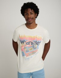 Κρέμα μπλουζάκι με πολύχρωμο γραφικό που περιλαμβάνει το κείμενο "Wrangler" και μια retro κασετόφωνο με ακουστικά, συνδυασμένο με ανοιχτό μπλε τζιν.