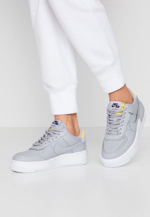 Harmaat Nike Air Force 1 -kengät sisältävät nahkapinnan, sileän tekstuurin, valkoisen tukevan pohjan sekä keltaisia ja violetin korostuksia kantapäässä ja kielekkeessä.