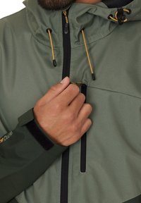 Green waterproof jacket featuring a zippered pocket, adjustable hood with yellow drawstrings, and black velcro wrist closures.
