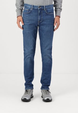 Jeans de mezclilla azul oscuro con un corte ajustado, diseño de cinco bolsillos y cierre de botón. Llevados con zapatillas grises.