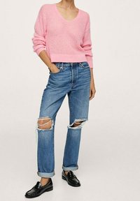 Maglione lavorato a maglia rosa chiaro con scollo a V, abbinato a jeans blu strappati con ginocchia lacerate e orli arrotolati. Mocassini neri completano il look.