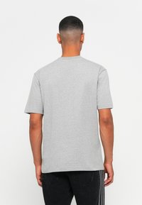 Muž s krátkymi vlasmi oblečený v jednoduchom šedom tričku s krátkym rukávom a čiernych nohaviciach, stojí chrbtom k bielej pozadí.