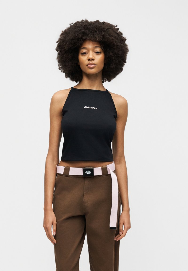 Woman with afro hairstyle wearing a black crop top with "Dickies" text, brown pants, and a pink belt against a plain gray background.
