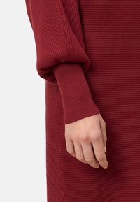 Rotes, geripptes Strickkleid mit einem lockeren, umgeschlagenen Ärmel. Der Stoff verfügt über ein strukturiertes Muster, das die warme Farbe und weiche Erscheinung betont.