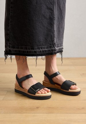 Zwarte leren sandalen met twee brede banden en een gespdetail. Kurken voetbed en zwarte rubberen sleehak. Rafelige zoom van een grijs denimrok zichtbaar.